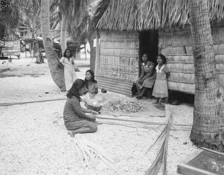 Women weaving palm branches outside their home, an example of household-based production