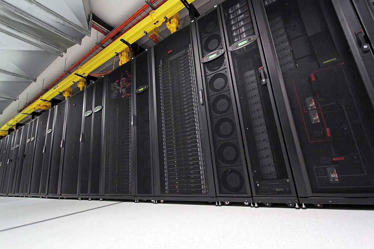 Rows of server racks inside a data centre, status lights glowing along dark cabinets with bundled cables running overhead