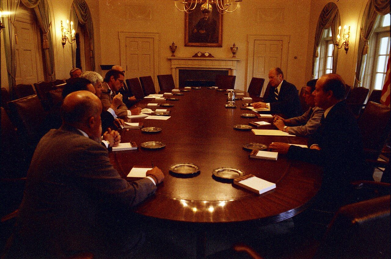 Advisers seated around a long conference table in a wood-panelled cabinet room, with a fireplace and framed portrait behind them