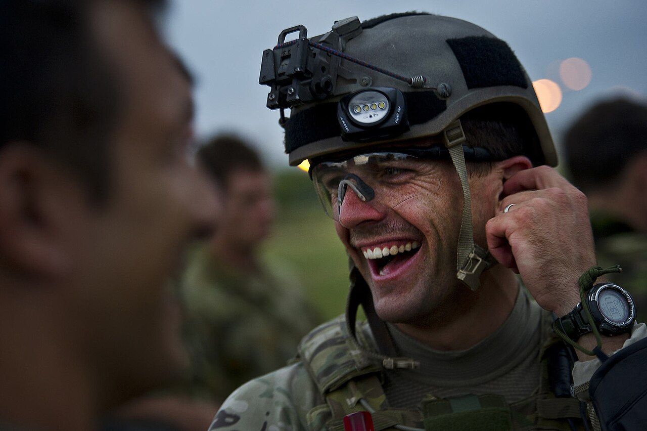 A service member in helmet and face paint laughing with colleagues standing beside him outdoors at dusk.